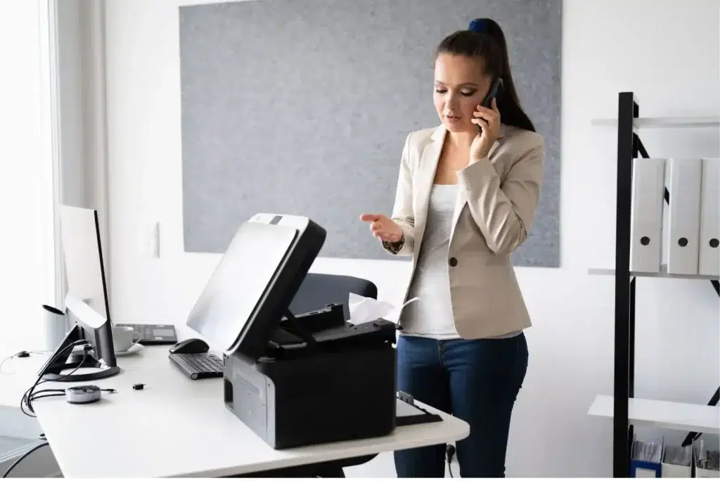 Profesional de oficina hablando por teléfono mientras revisa una impresora multifunción.